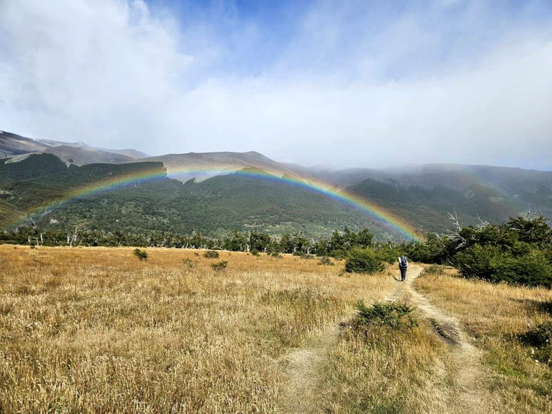 Un arc-en-ciel se déploie au-dessus de collines boisées tandis qu'une personne en randonnée suit un chemin de terre à travers une prairie dorée, en route vers le camping de Serón.