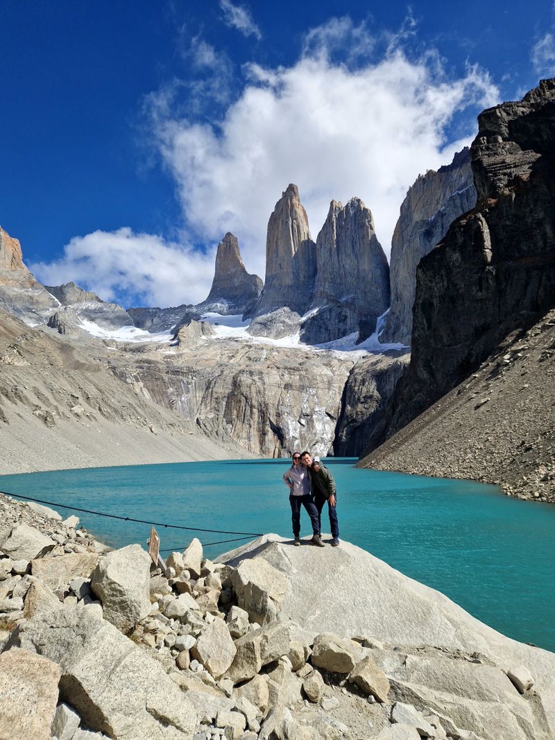 Au mirador Base Torres, deux personnes se tiennent sur un rocher au bord du lac turquoise, au pied de falaises striées de neige et des trois tours de granit.
