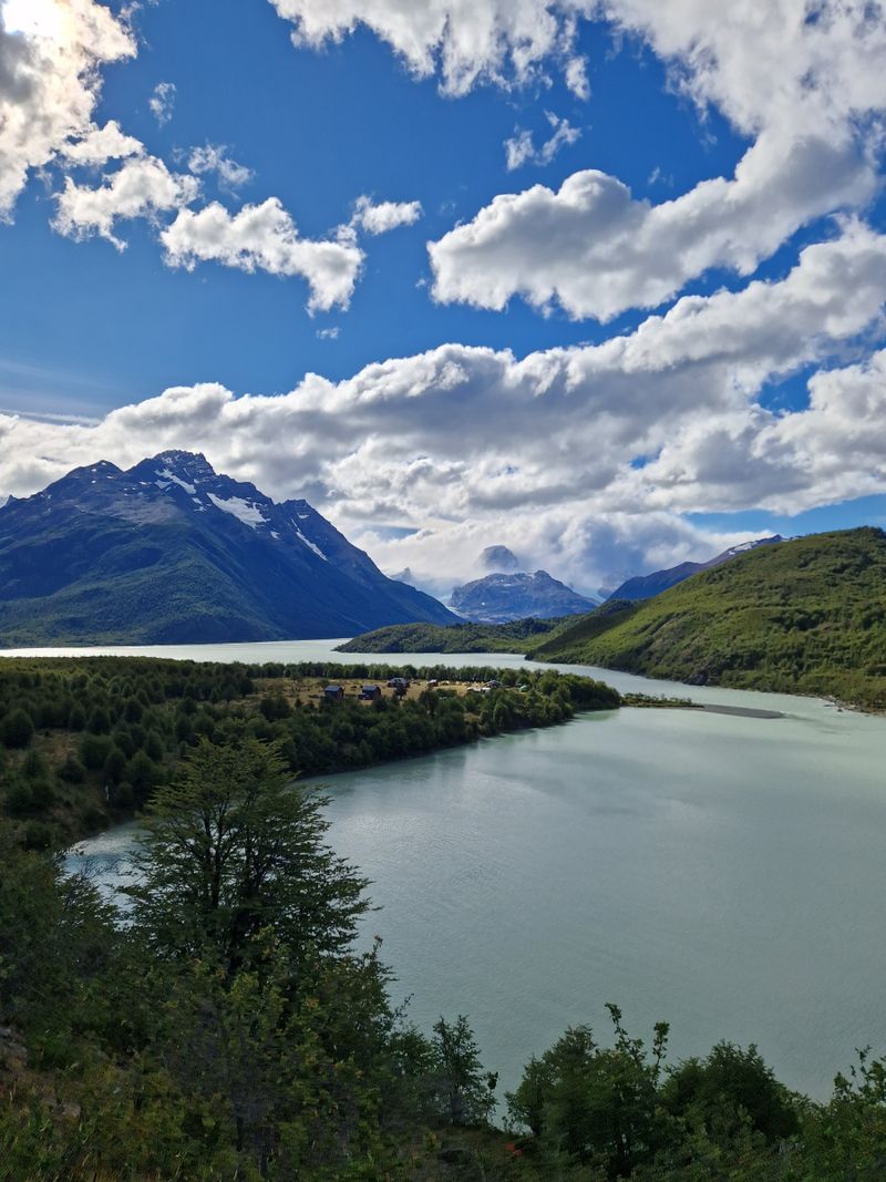 Le lac Dickson, d'un vert pâle, s'incurve le long d'une rive boisée où se trouvent les bâtiments du camping Dickson, sous des sommets sombres et de gros nuages blancs.