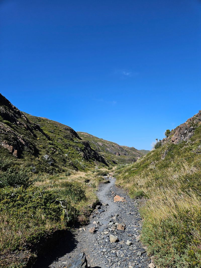 Un sendero pedregoso sube por un estrecho valle herboso entre laderas cubiertas de matorral y afloramientos rocosos, bajo un cielo azul intenso y despejado.