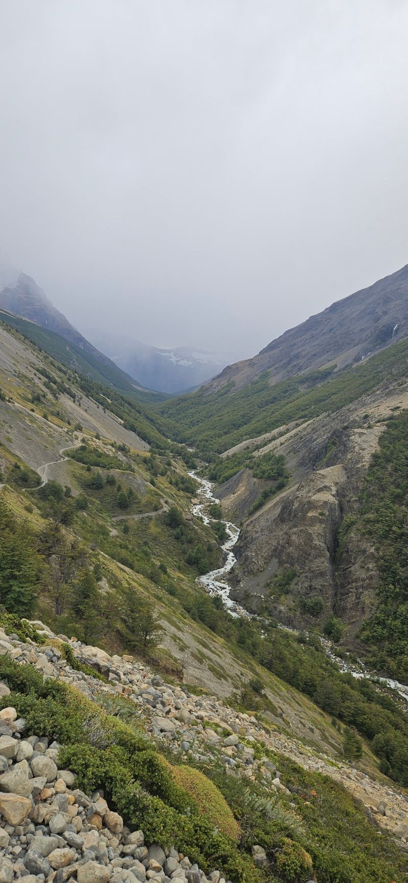 Un sendero sube por una ladera pedregosa sobre un río de deshielo que serpentea por un valle verde y encajonado. Las nubes bajas ocultan las cumbres.