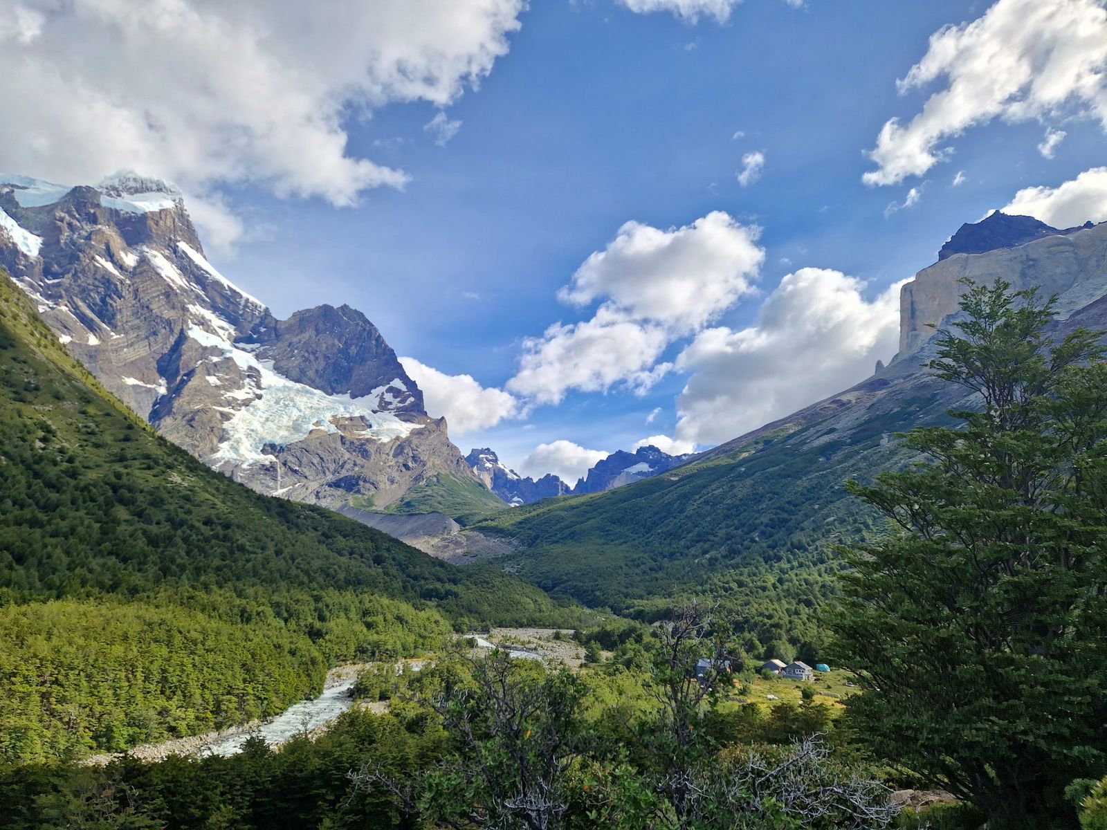 El glaciar colgante del Paine Grande se alza sobre el verde valle del Francés, donde un río de deshielo serpentea por el bosque junto a pequeñas construcciones.